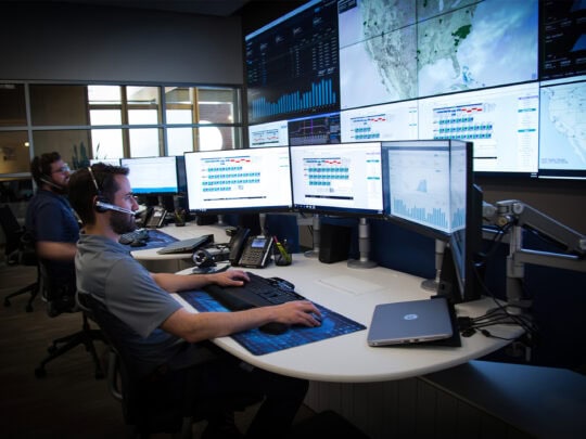 A man sits at a desk facing a computer with four monitors, in front of a wall of computer screens showing data, behind him another man does the same