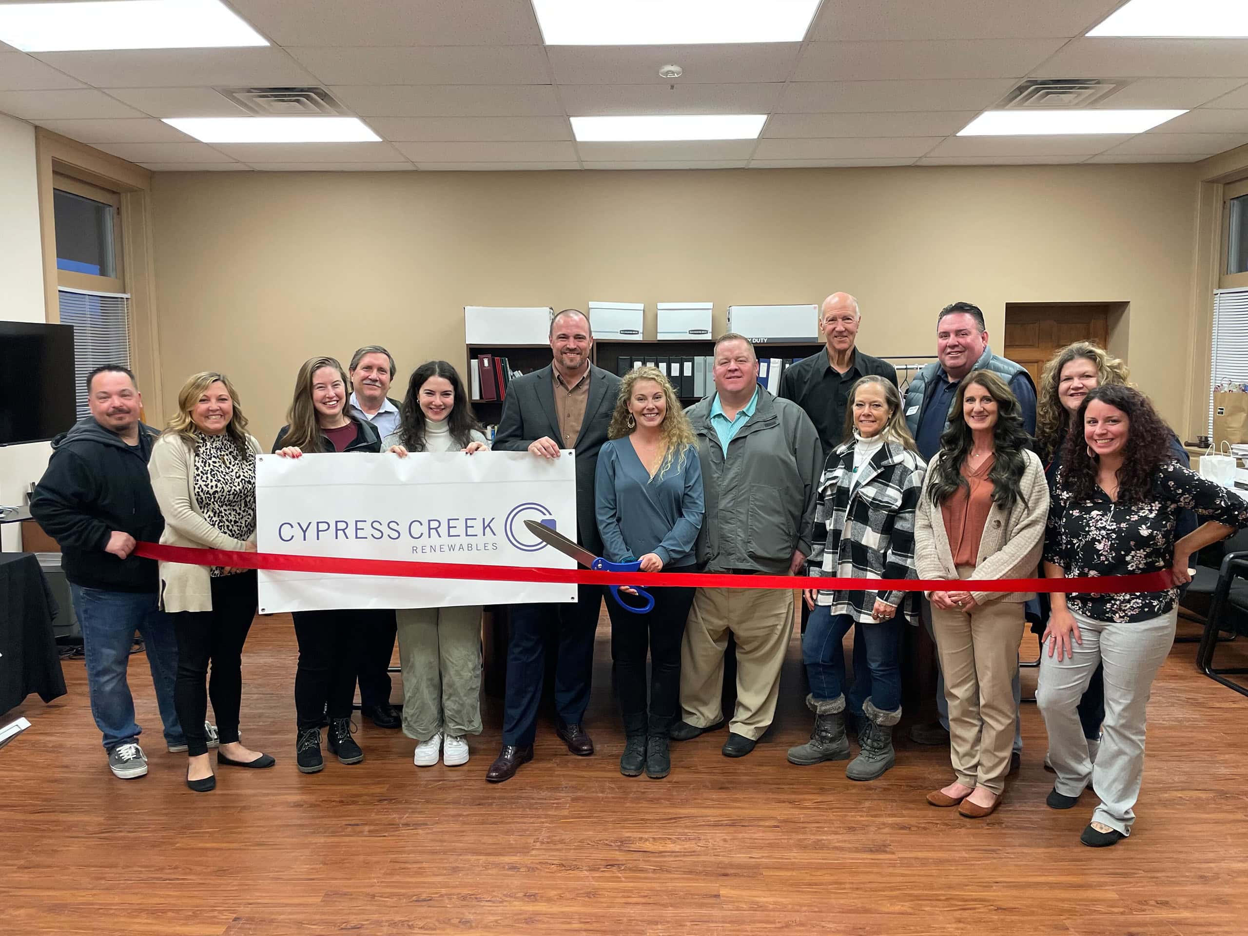 A group of 14 people pose in a line behind a red ribbon as a woman in the middle holds large scissors. Three people hold a Cypress Creek Renewables banner.