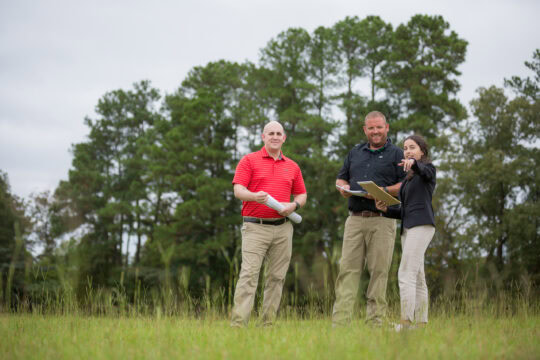 A group of three people stand talking in a field. A woman points at something.