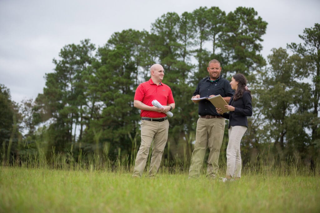 Three people stand in a field holding paperwork and talking