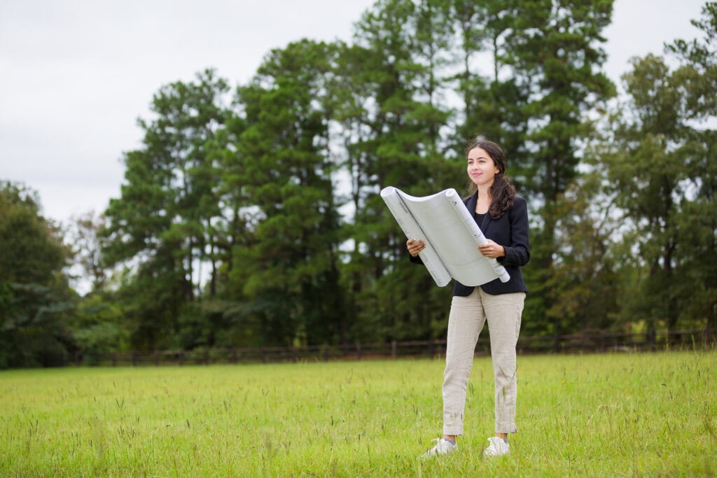 A young woman looks over a set of blueprints in an empty field.