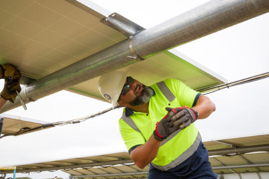 A man wearing safety gear looks under a solar panel as it is being installed.