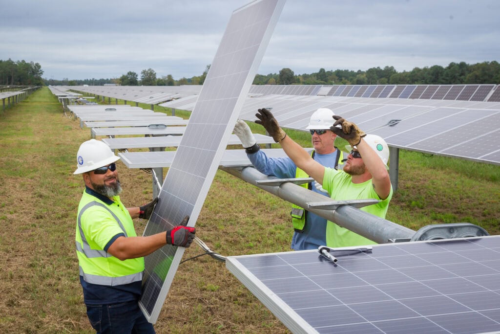 One man in a hard hat holds a solar panel as two men reach of the other end of it among a row of solar panels