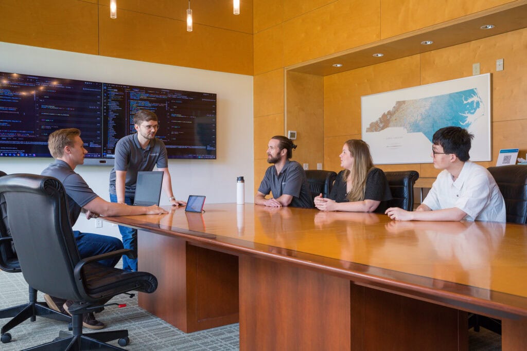 A man stands at the head of a conference table with data on televisions behind him as people around the table listen to him