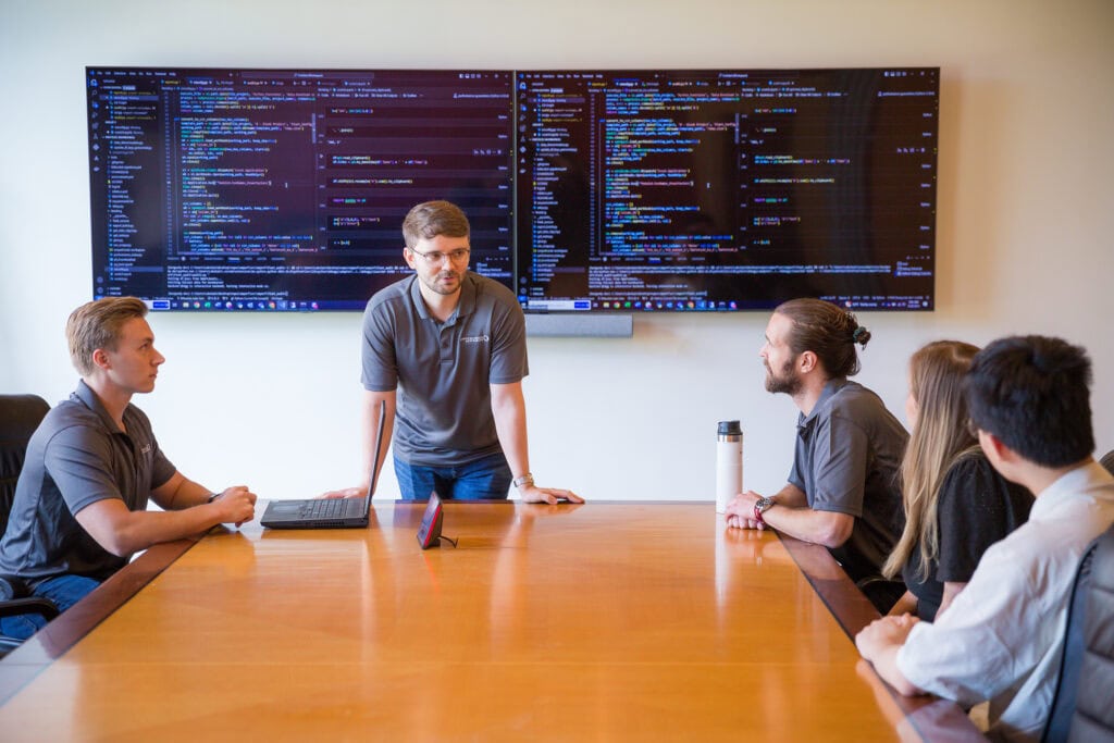 A man stands at the head of a conference table with data on televisions behind him as people around the table listen to him