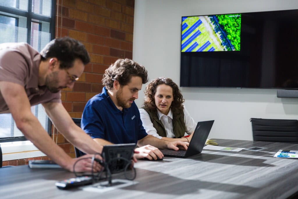 At a table one man looks at a piece of computer equipment as another man and a woman look at a laptop screen