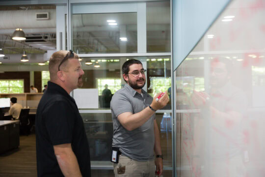 Two men stand at a whiteboard, one holds a marker and talks to the other about something written on the board