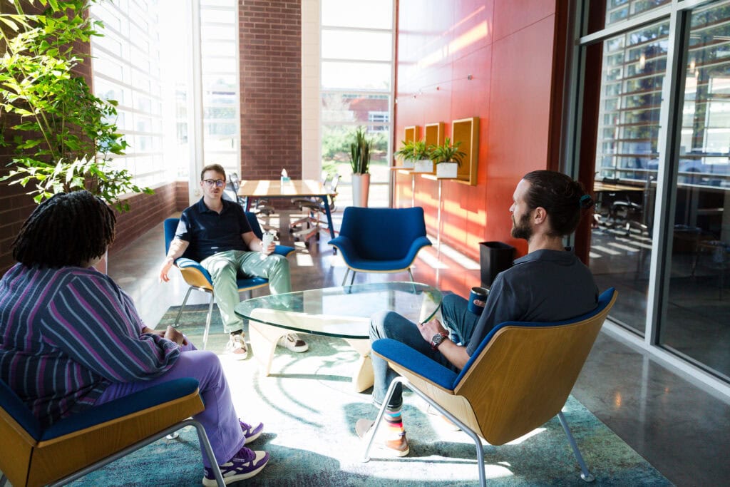 Three people sit in blue chairs around a glass table in a sunlit meeting space