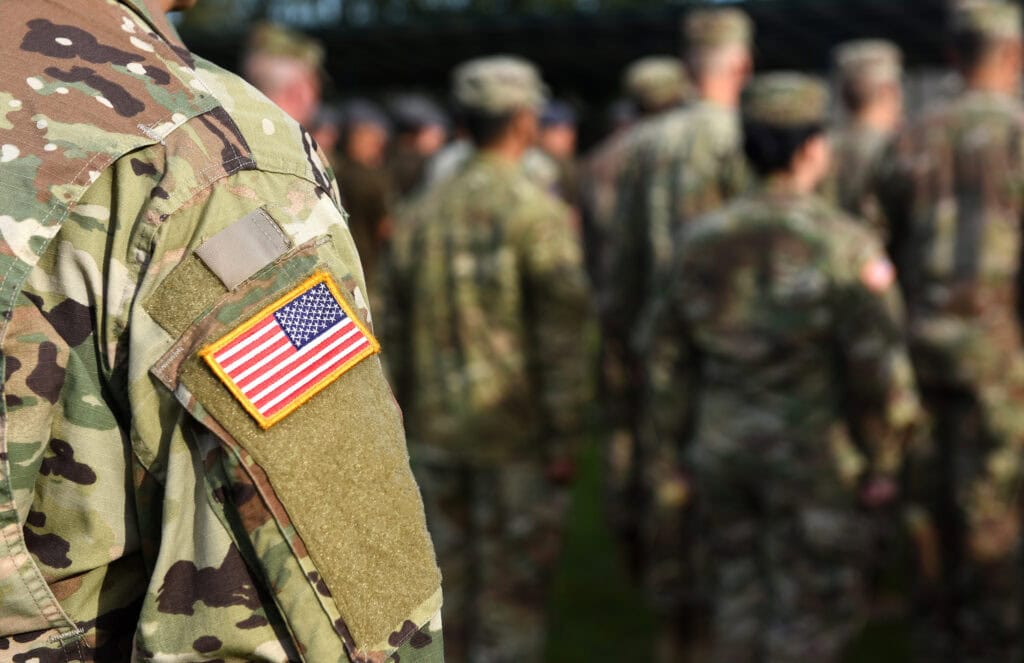 An American flag patch is shown on the shoulder of a camouflage uniform in focus on the right as soldiers in similar uniforms are show out of focus on the right.