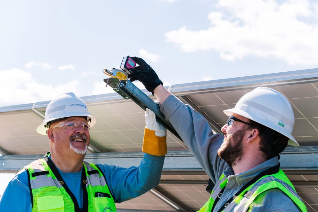 Two men in safety vests and hard hats use a device to take a measurement on the back of a solar panel