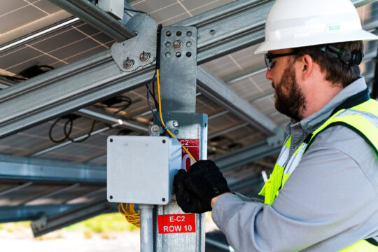 A man in safety gear works with wires under a solar panel