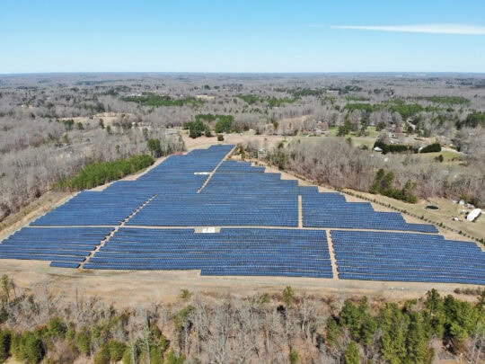 An aerial view of a solar farm in an early winter landscape under a pale blue sky