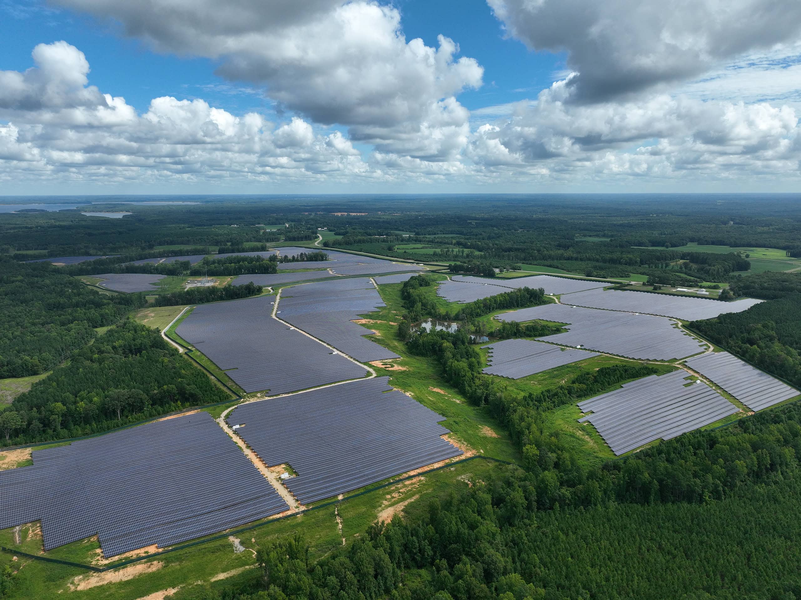 A solar farm is seen from above surrounded by trees with a small pond in the center.