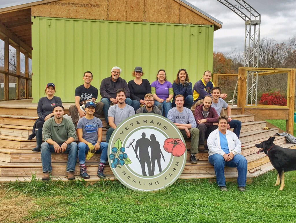 A group of 15 people pose on the steps of a deck with a large sign reading "Veterans Healing Farm"