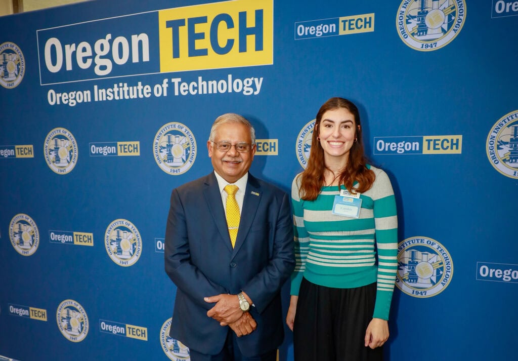 A man and a woman pose in front of a backdrop with an Oregon Tech logo on it.