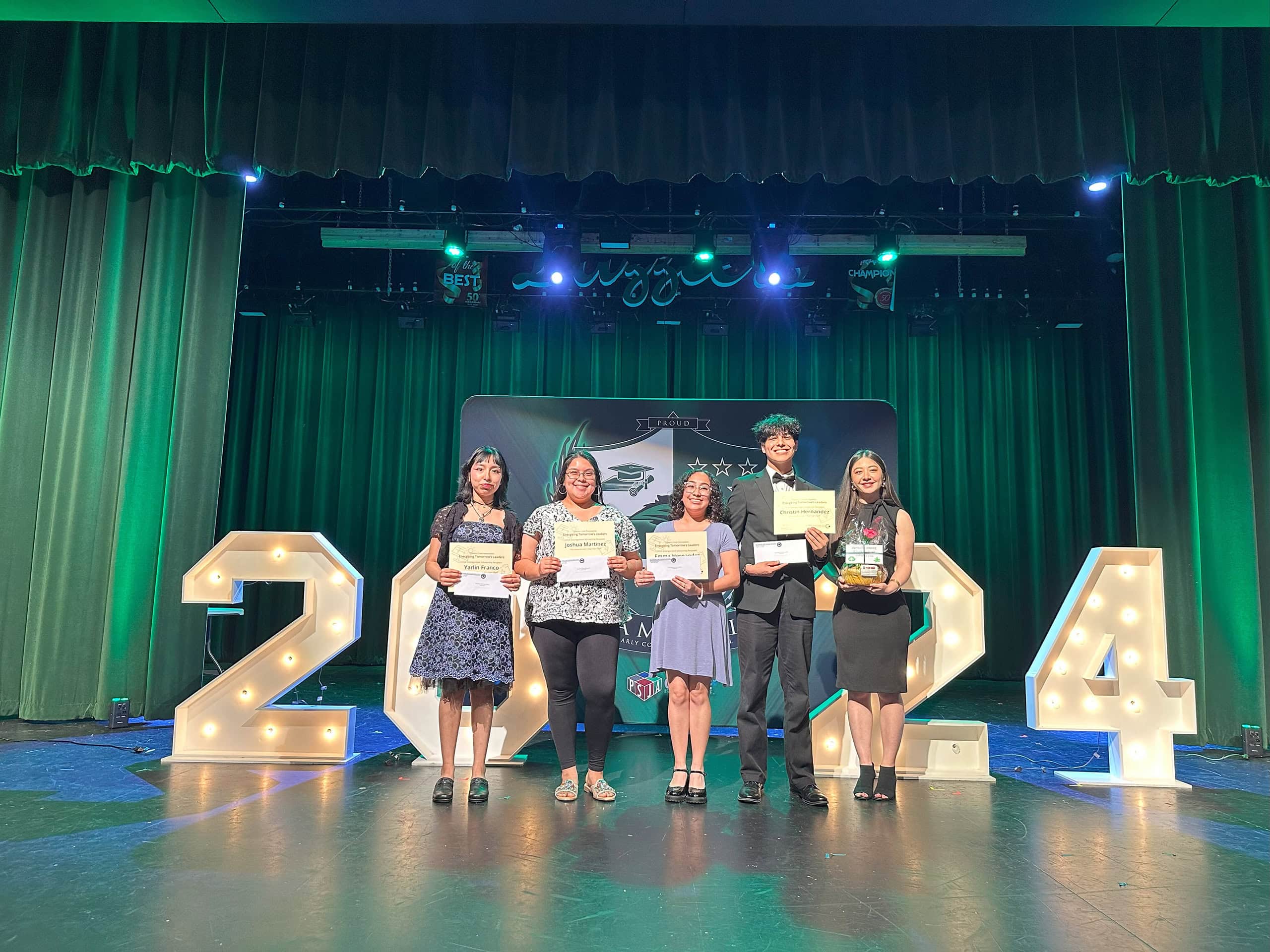 A group of five teenagers stand in a line on a stage holding certificates in front of large lit up numbers that read "2024"