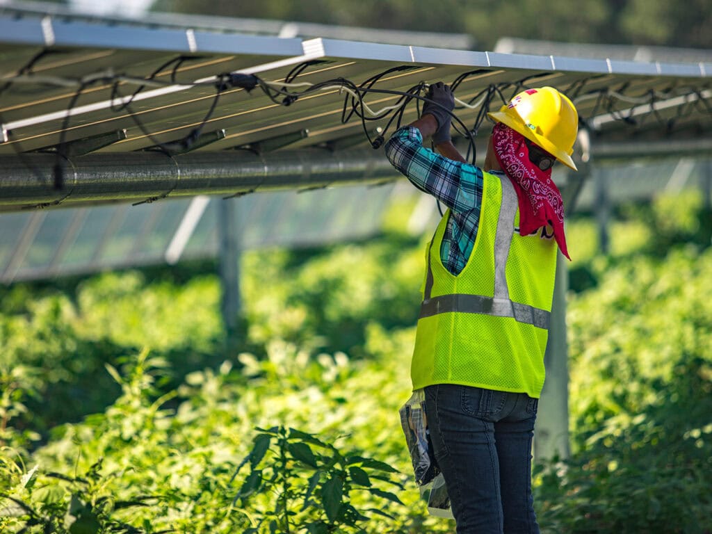 A worker in yellow safety gear works with wires under a solar panel in a grassy field