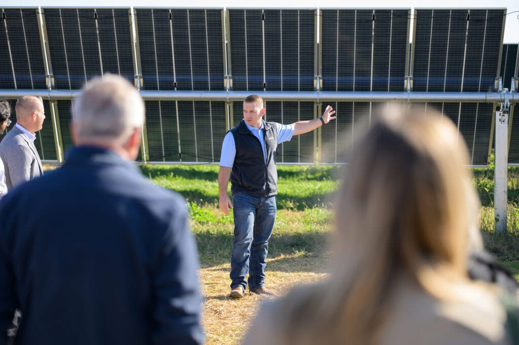 A man seen through out of focus people in the foreground gestures to a row of solar panels