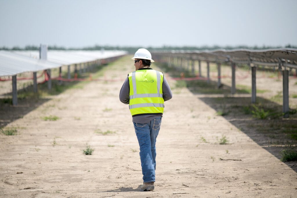 A man in a safety vest and hard hat walks between rows of solar panels in a desert