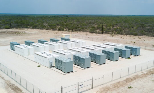 Large battery storage shipping containers are seen in rows surrounded by a fence in a desert and shrub landscape with a grey blue sky