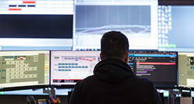 A man sits at a computer surrounded by monitors with various displays on them