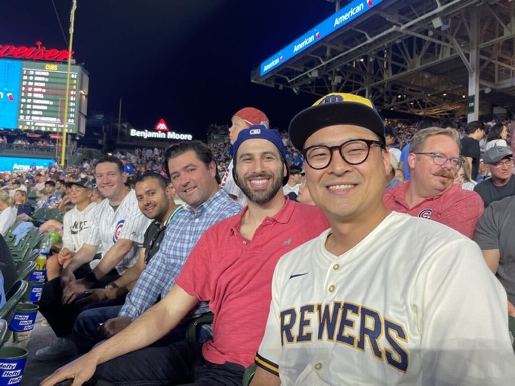 A group of men smile at the camera while sitting in the stands at a baseball game