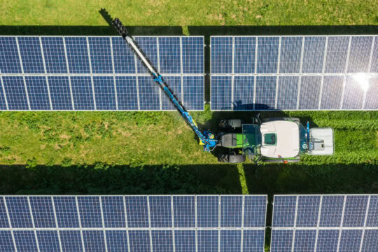 A tractor with a large arm squirts water onto a row of solar panels as it drives between two rows