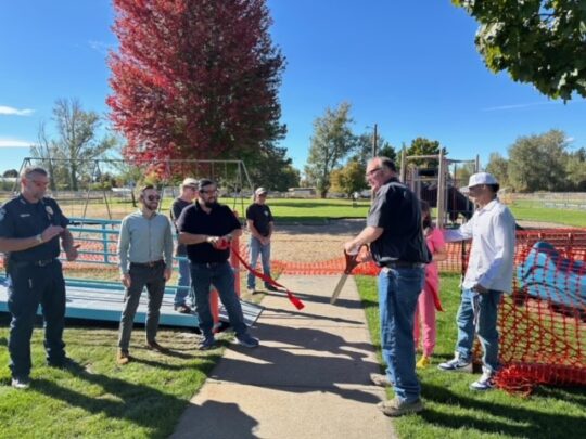 A red ribbon is cut by large scissors in front of a playground as people look on